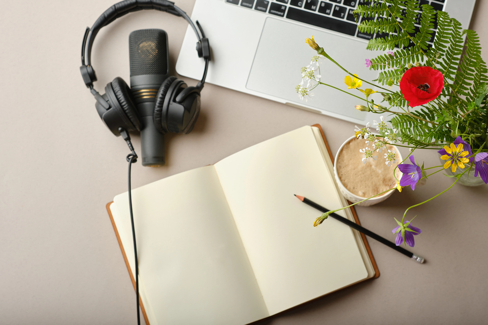 An open notebook with a pencil on it sits beside a cup of coffee, a laptop, a microphone with headphones, and a vase of colourful flowers on a light desk.