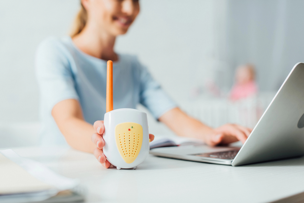 A woman sitting at a desk with a laptop, holding a baby monitor in her hand. The background is softly blurred.