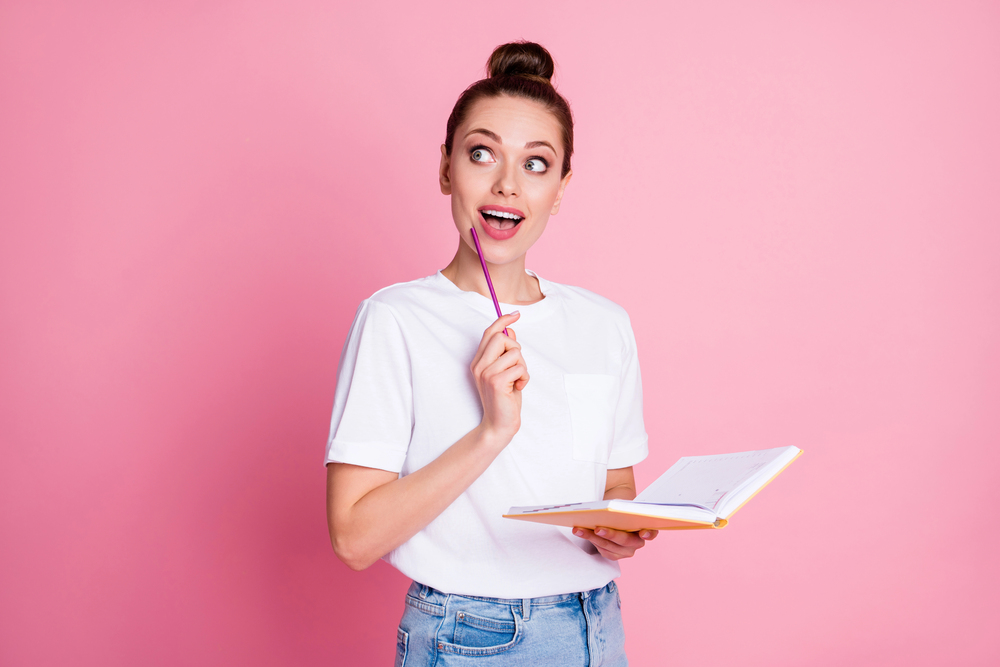 A young woman in a white T-shirt holds an open notebook and a pencil, looking thoughtful with a slight smile, standing against a pink background.