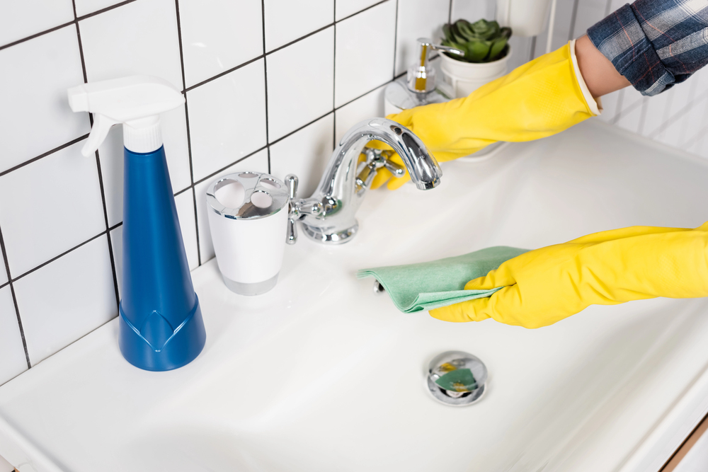 Person wearing yellow rubber gloves cleaning a bathroom sink and tap with a green cloth; a blue spray bottle and toothbrush holder are nearby, with white tiled walls in the background.