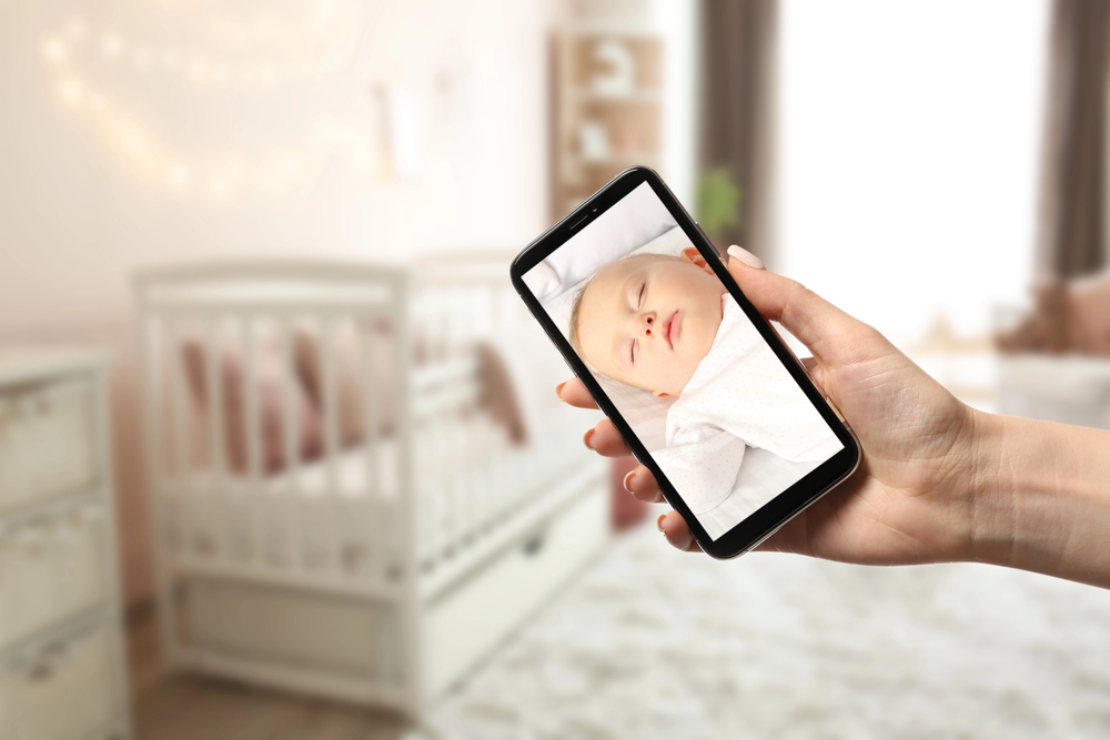 A hand holds a mobile phone displaying a live video of a sleeping baby, with a blurred nursery and cot visible in the background.