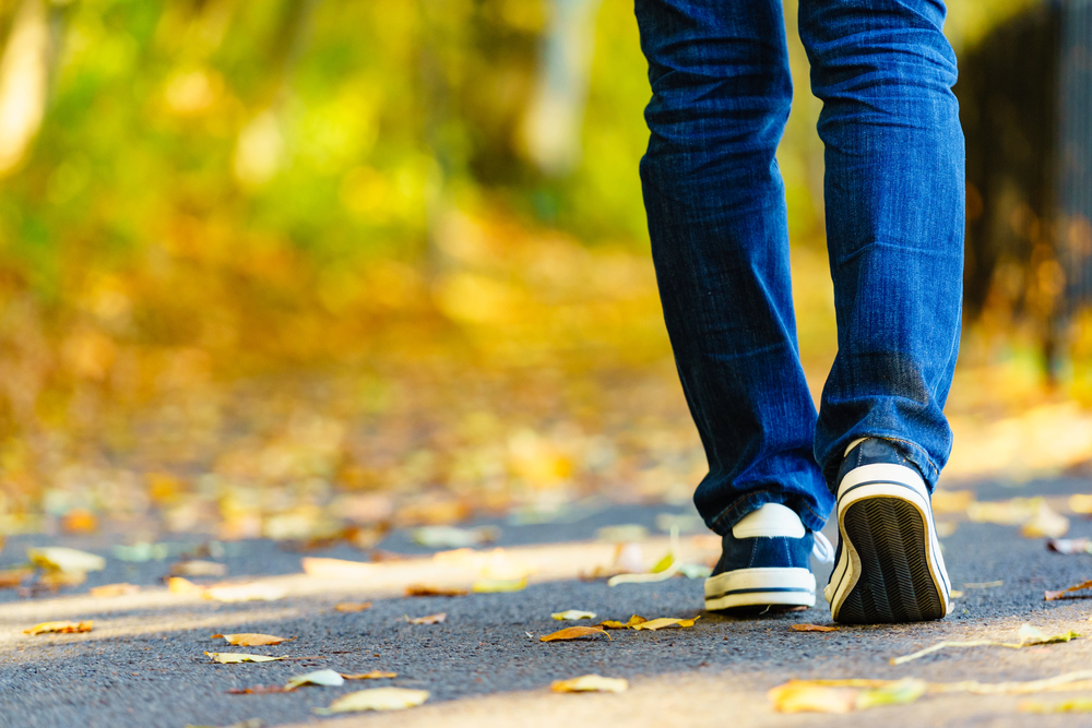 A person wearing blue jeans and black trainers walks on a path covered with scattered yellow leaves, surrounded by blurred greenery and sunlight.