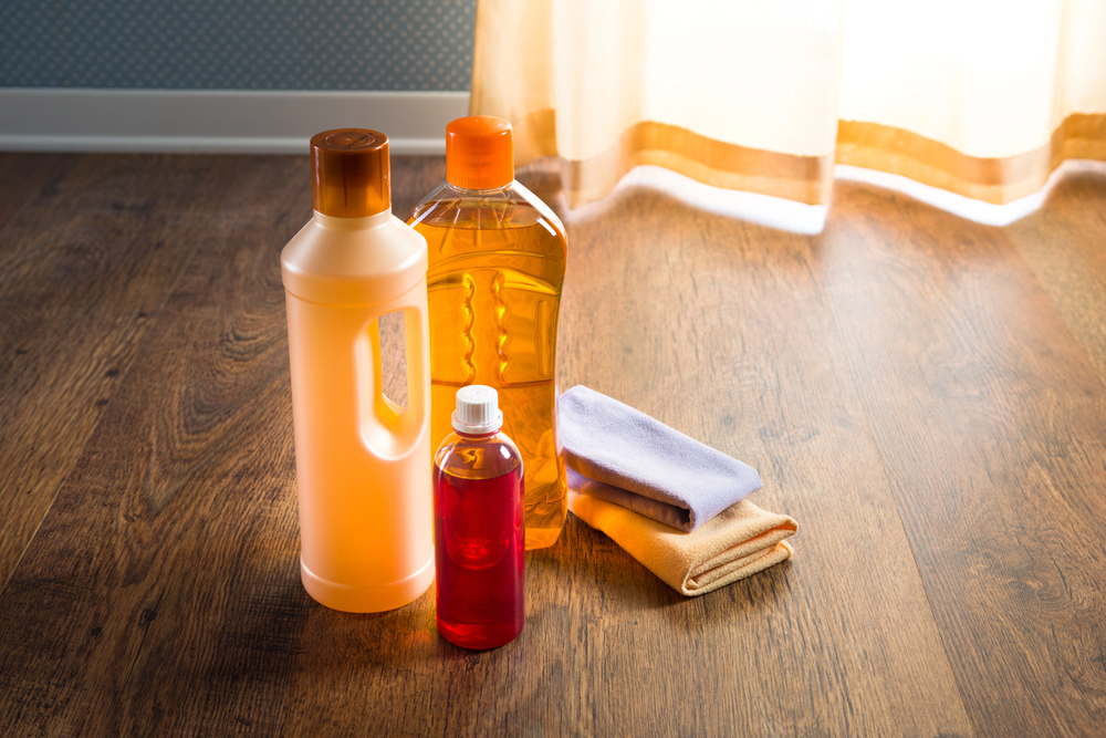 Three plastic bottles with cleaning liquids and two folded microfibre cloths are placed on a wooden floor near a window with sheer curtains and sunlight streaming in.