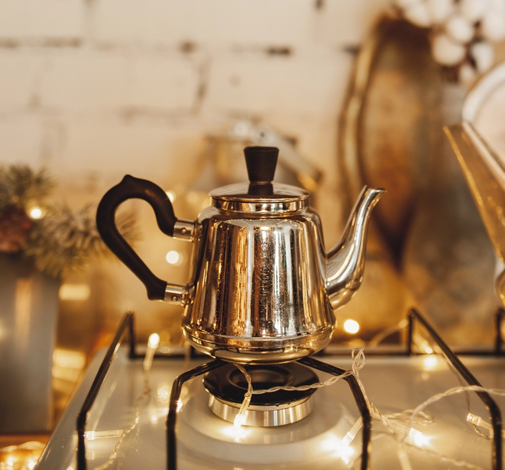A shiny metal teapot with a black handle and lid sits on a gas hob burner, surrounded by warm, glowing fairy lights in a cosy, festive kitchen setting.