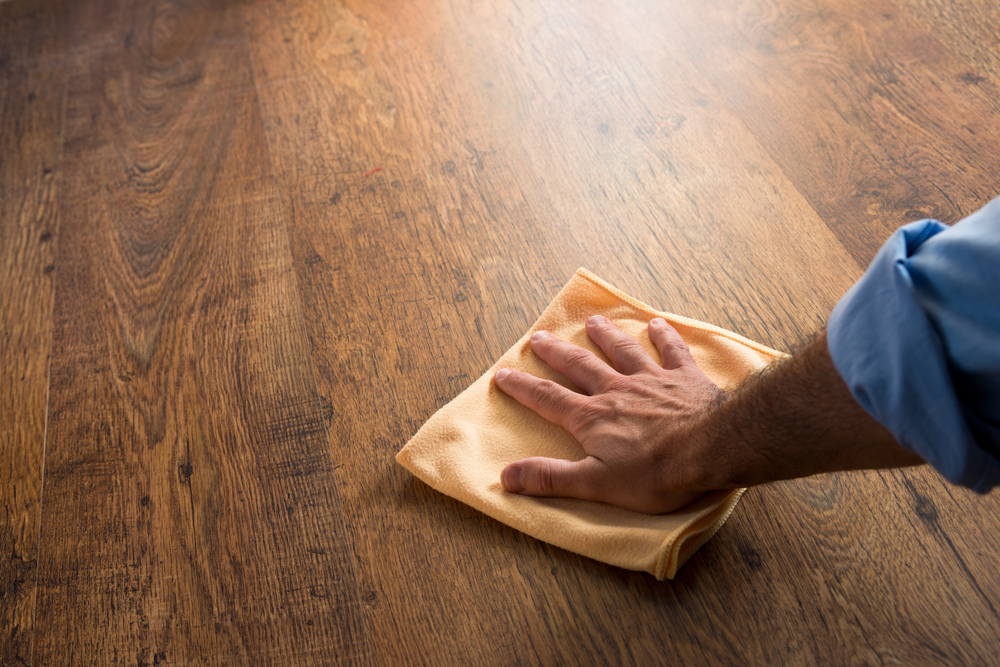 A person’s hand wipes a wooden table with a yellow cloth, cleaning the surface. The person is wearing a blue long-sleeved shirt. The table has a natural brown wood grain pattern.