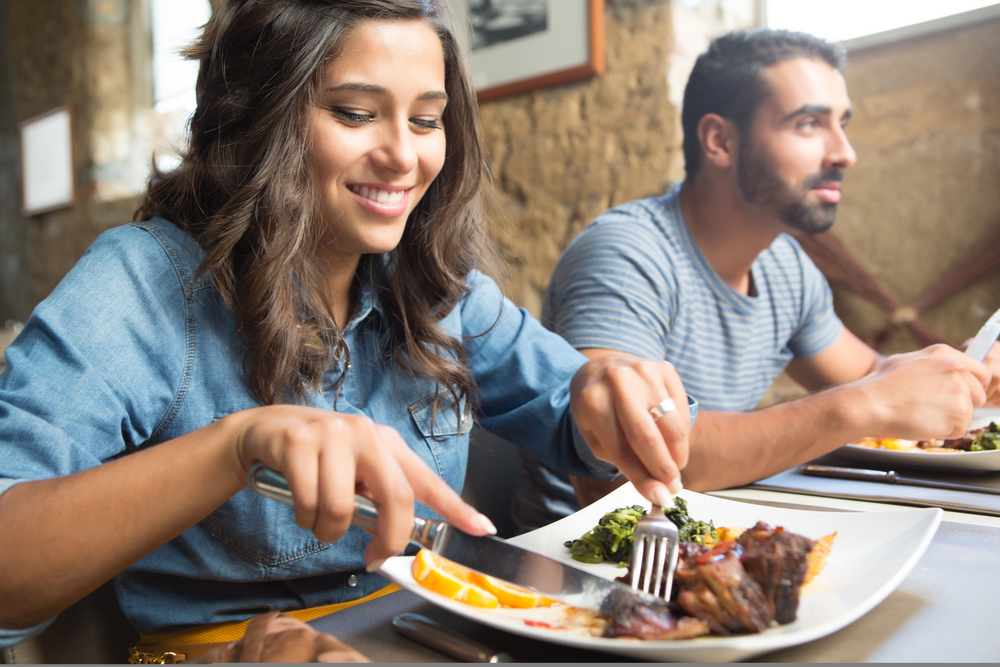 A woman smiles while cutting food on her plate at a restaurant, with a man sitting beside her. Both appear to be enjoying their meal.