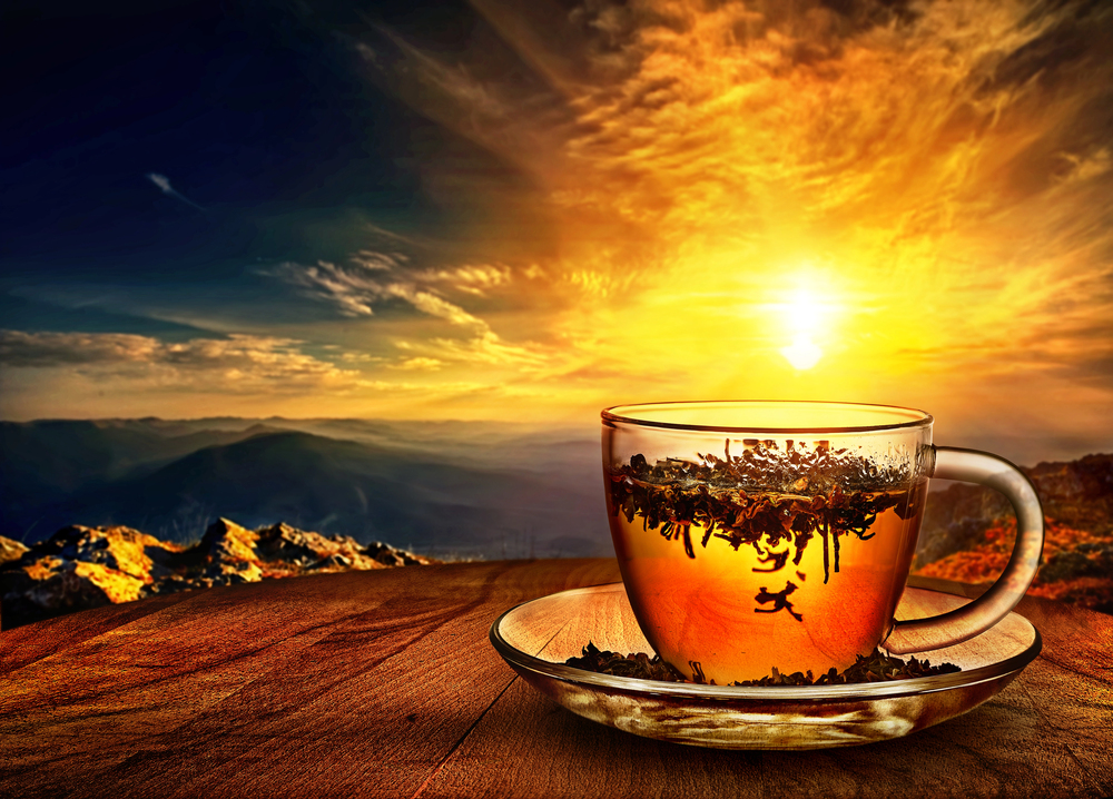 A glass cup of tea with floating tea leaves sits on a wooden surface, with a scenic view of mountains and a vibrant golden sunset in the background.