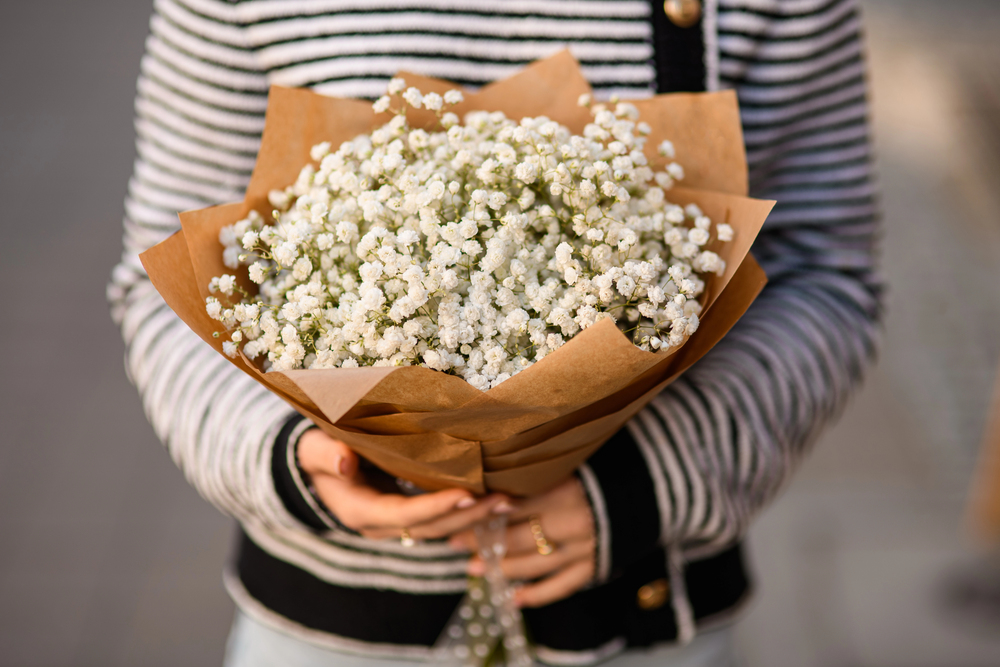 A person in a striped jumper holds a bouquet of small white flowers wrapped in brown paper. The flowers appear to be baby's breath. The background is blurred.