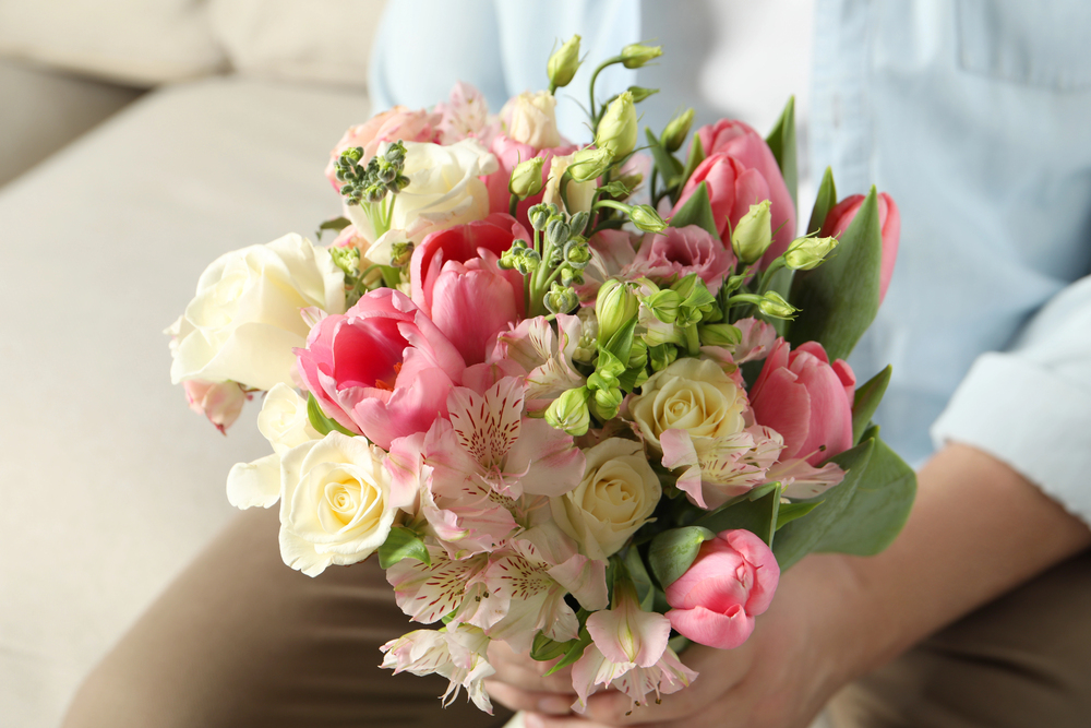 A person holding a bouquet of pink tulips, white roses, and pale pink flowers, seated on a light-coloured sofa and wearing tan trousers and a light blue shirt.