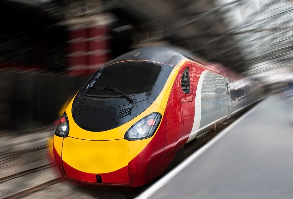 A modern, red and yellow high-speed train travels quickly along tracks at a station, with motion blur emphasising its speed and sleek design.
