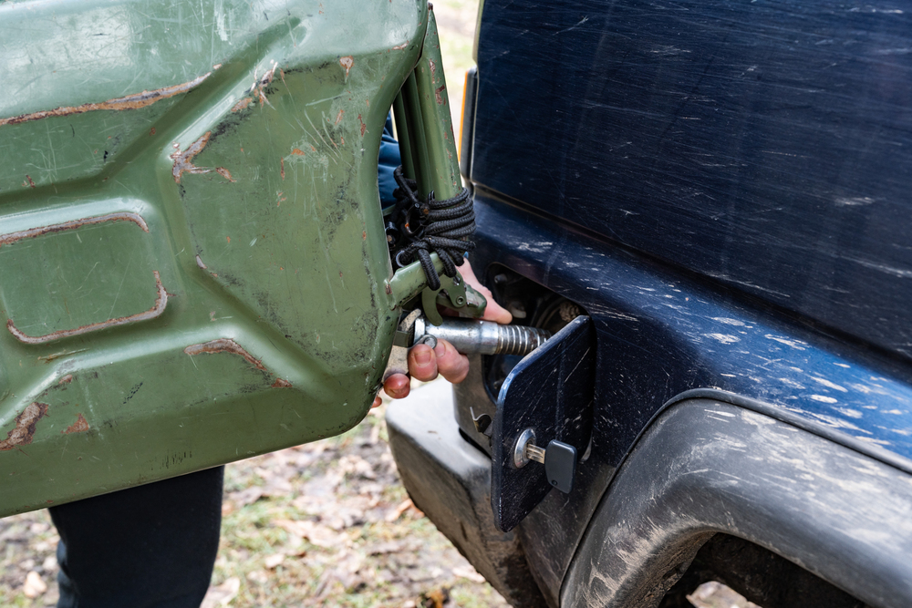 A person is pouring fuel from a green metal jerry can into the petrol tank of a dark blue vehicle. The scene appears to be outdoors, and the vehicle is slightly dirty.