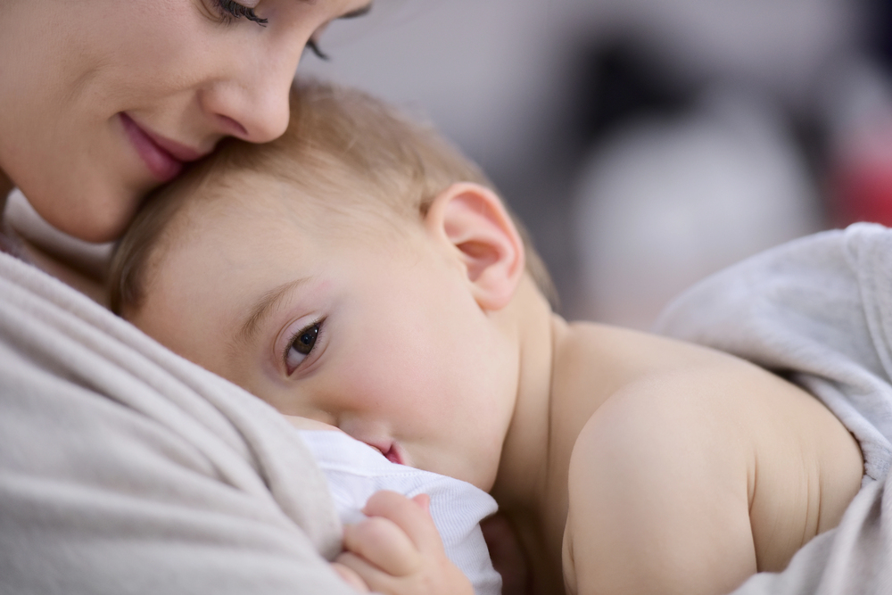 A close-up of a mother lovingly nursing her baby, who is cuddled against her chest and looking outward while breastfeeding. Both appear calm and content, wrapped in a soft, cosy blanket.