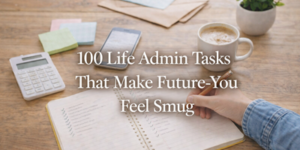 A person writes in a notebook on a wooden desk with a coffee cup, calculator, sticky notes, and phone nearby. Overlaid text reads: 100 Life Admin Tasks That Make Future-You Feel Smug.