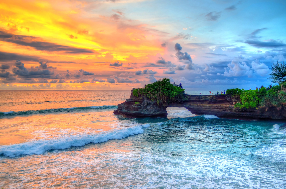 Colourful sunset over the ocean at Tanah Lot, Bali, with dramatic clouds, vibrant orange and blue sky, waves, and a rocky outcrop covered in green plants extending into the water.
