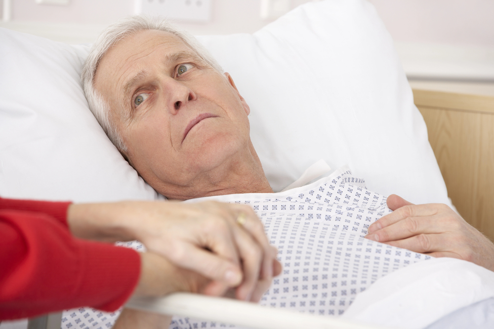 An elderly man lies in a hospital bed, looking thoughtfully to the side. He wears a hospital gown and rests one hand on his chest. Another person gently holds his other hand in a comforting gesture.