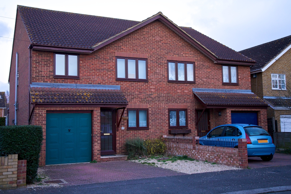 A semi-detached red-brick house with brown roof tiles, two front doors, and two garages. A blue car is parked on the drive to the right, and bushes are beside the houses.