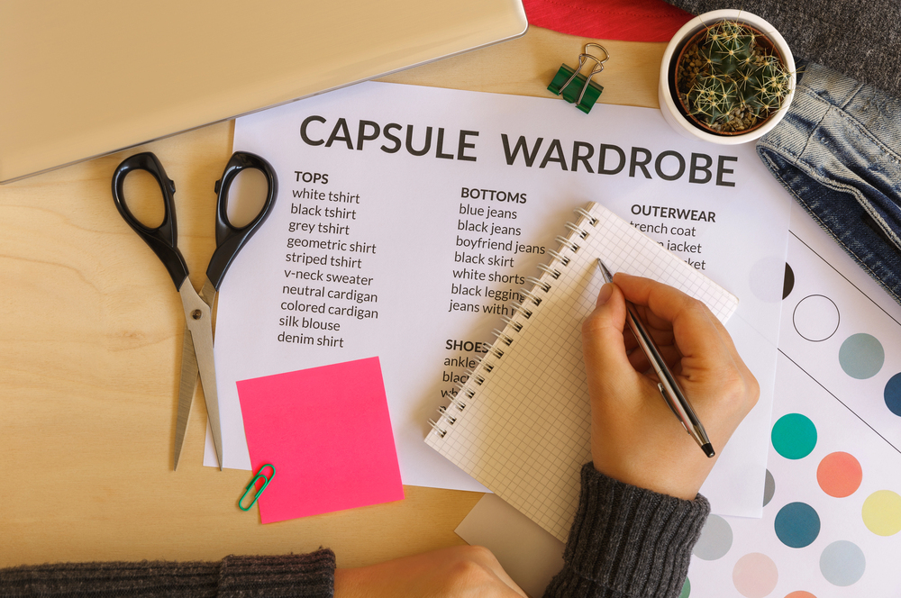 A person writes in a notepad next to a Capsule Wardrobe checklist on a desk with scissors, sticky notes, a plant, paper clips, and fabric swatches.