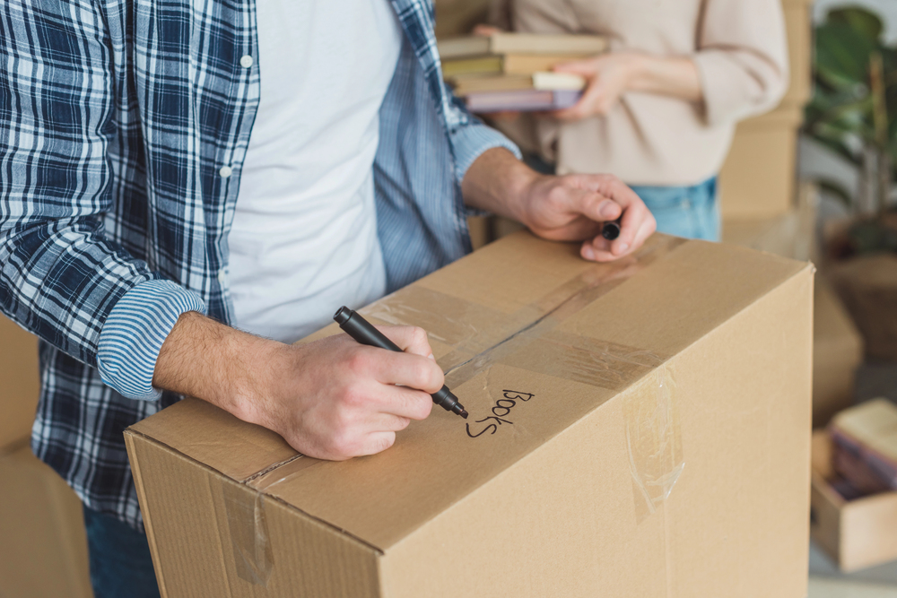 A person in a checked shirt labels a cardboard box with the word “Books” using a black marker, while another person in the background holds several books.
