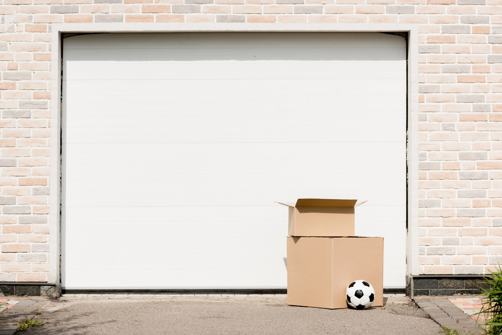 Two cardboard boxes, one open, and a football are placed in front of a closed white garage door set in a light brick wall.