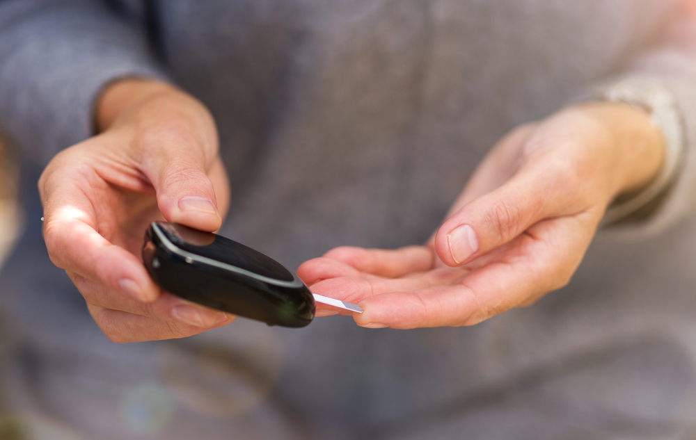 A person uses a blood glucose metre to check their blood sugar level, holding the device in one hand and applying a small drop of blood from a fingertip in the other hand.