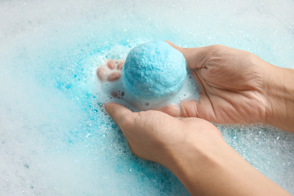 Two hands hold a blue bath bomb in foamy, soapy water, with blue colour spreading from the bath bomb into the bubbles.