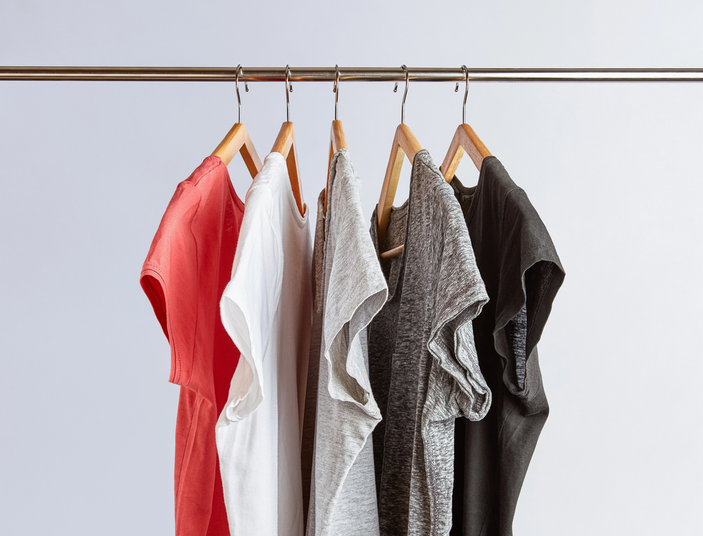 Five short-sleeve T-shirts in red, white, light grey, dark grey, and black hang neatly on wooden hangers from a metal clothes rail against a plain, light background.