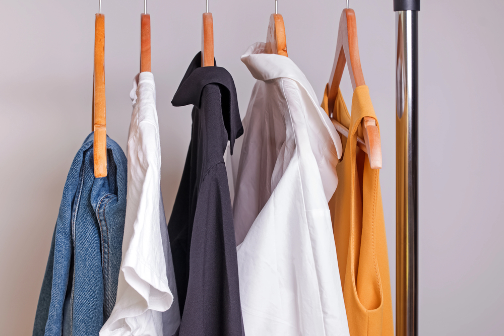 Five shirts hang on wooden hangers on a metal clothes rail. The shirts are blue denim, white, black, white, and mustard yellow, arranged from left to right, against a plain background.
