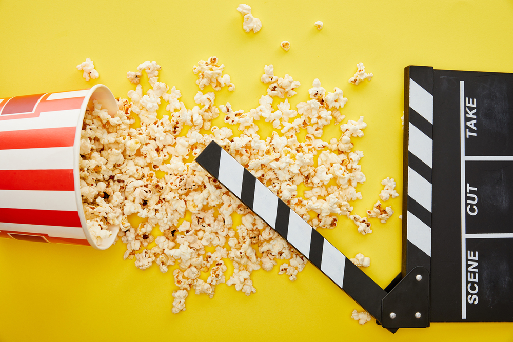 A tipped-over striped popcorn bucket spills popcorn onto a yellow surface next to a black and white film clapperboard.