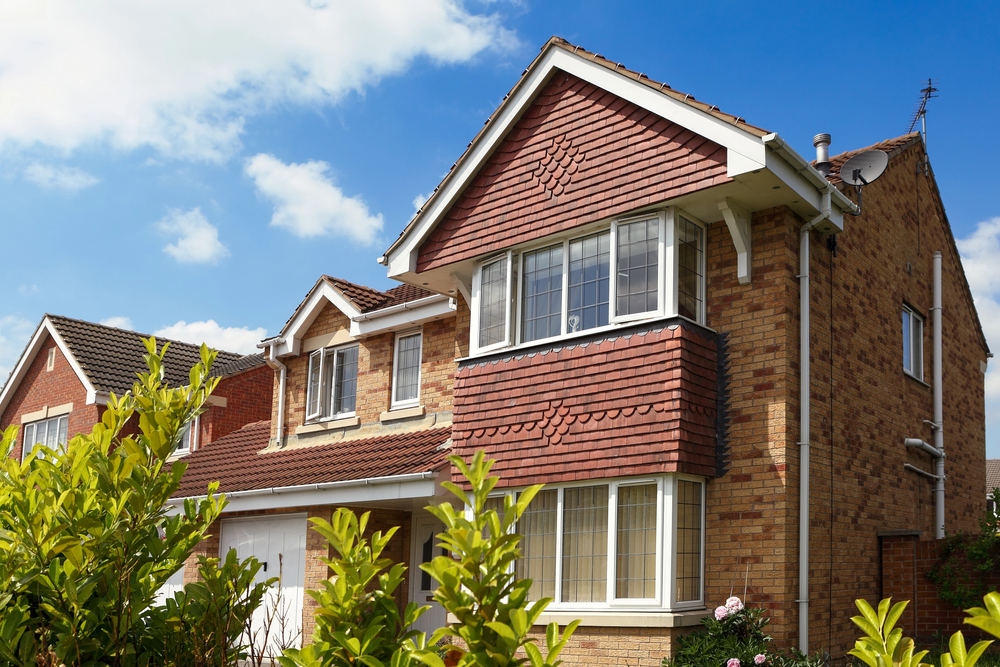 A two-storey brick house with red embellished cladding, white trim, large windows, and a double garage, surrounded by greenery, under a partly cloudy sky.