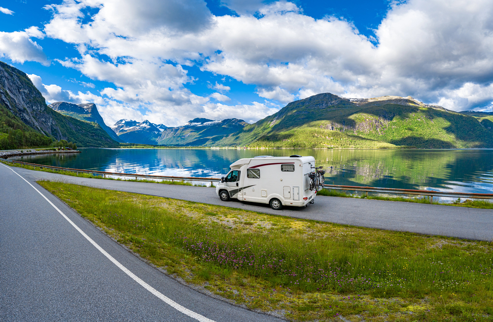 A white camper van parked beside a scenic lake, surrounded by green mountains and cloudy blue skies, with its reflection visible in the calm water. A road runs alongside the van and the lake.