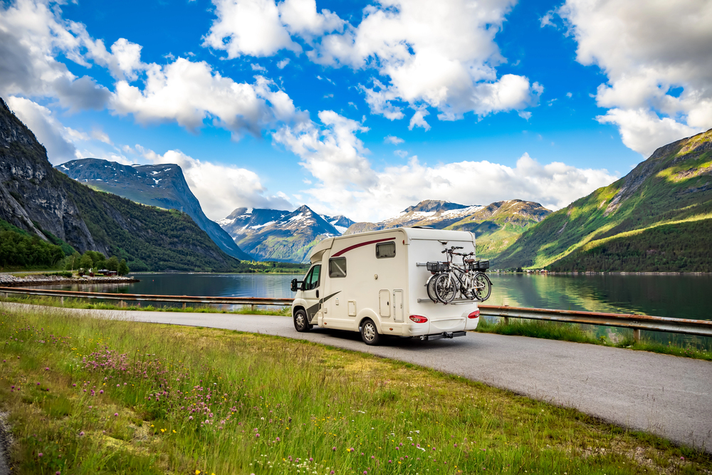 A white camper van with bicycles on the back is parked by a calm mountain lake, surrounded by green grass, mountains, and a partly cloudy blue sky.