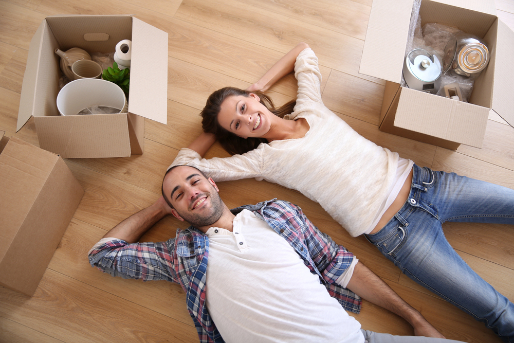 A smiling couple lies on a wooden floor, surrounded by open cardboard boxes filled with household items, suggesting they are in the process of moving into a new home.