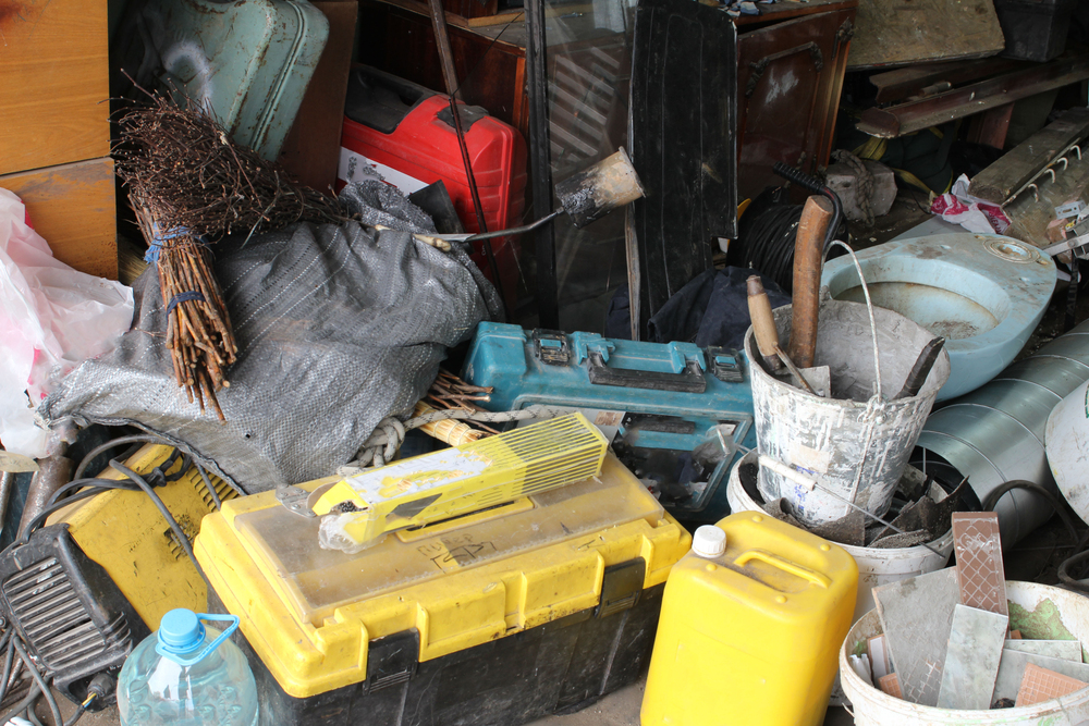 A cluttered pile of tools and equipment, including yellow and blue toolboxes, buckets with paint brushes, a water container, a bundle of sticks, and various building materials.