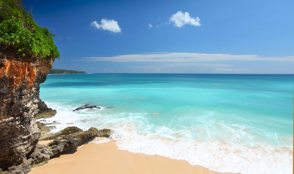A sandy beach with turquoise waves gently washing ashore, bordered by rocky cliffs covered in green vegetation under a bright blue sky with scattered clouds.