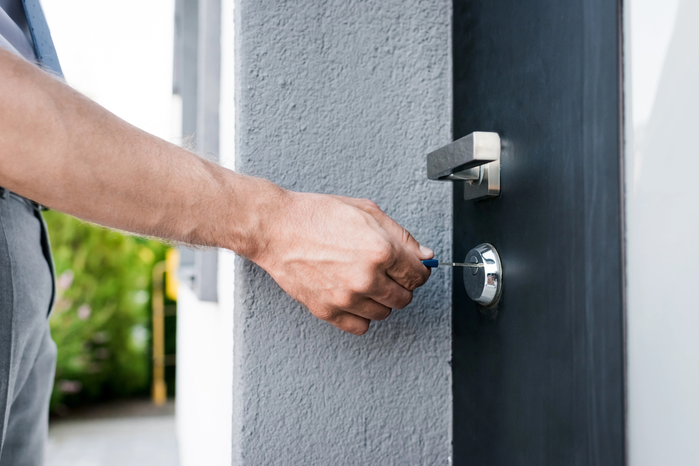 A person’s hand is inserting a key into the lock of a modern black door with a silver handle. The background includes a grey wall and some greenery outside.