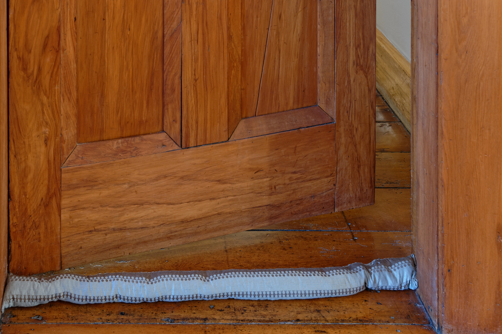 A wooden door is slightly open, with a fabric draught stopper placed at the bottom to block the gap. The floor and door frame are also wooden.