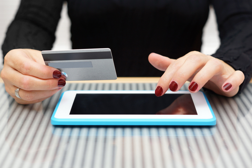 A person with red painted nails holds a credit card in one hand and uses the other hand to touch a tablet with a blue case on a striped glass table.