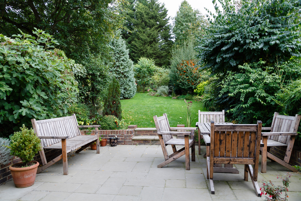 A stone patio with wooden benches, chairs, and a table overlooks a lush green garden with dense trees, bushes, and a manicured lawn. Potted plants line the patio, enhancing the natural, serene setting.