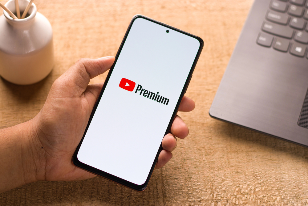 A hand holding a smartphone displaying the YouTube Premium logo on the screen, next to a laptop and a small white vase on a wooden surface.