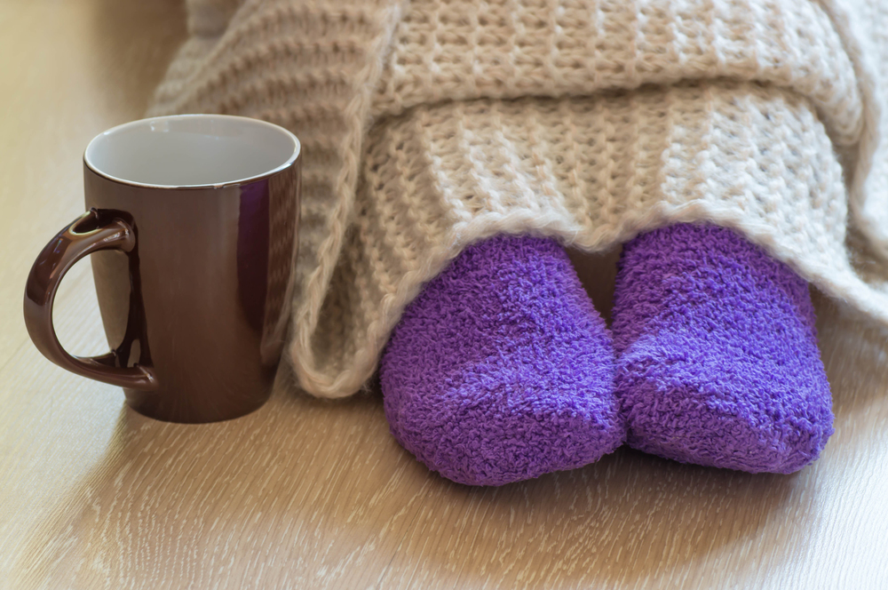 A person wearing purple fluffy socks sits on a wooden floor, legs covered with a beige knitted blanket. A brown mug is placed nearby, creating a cosy and warm atmosphere.
