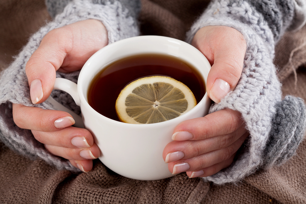 A person wearing cosy grey knitted sleeves holds a white mug filled with tea and a slice of lemon on top, resting on a brown textured blanket.