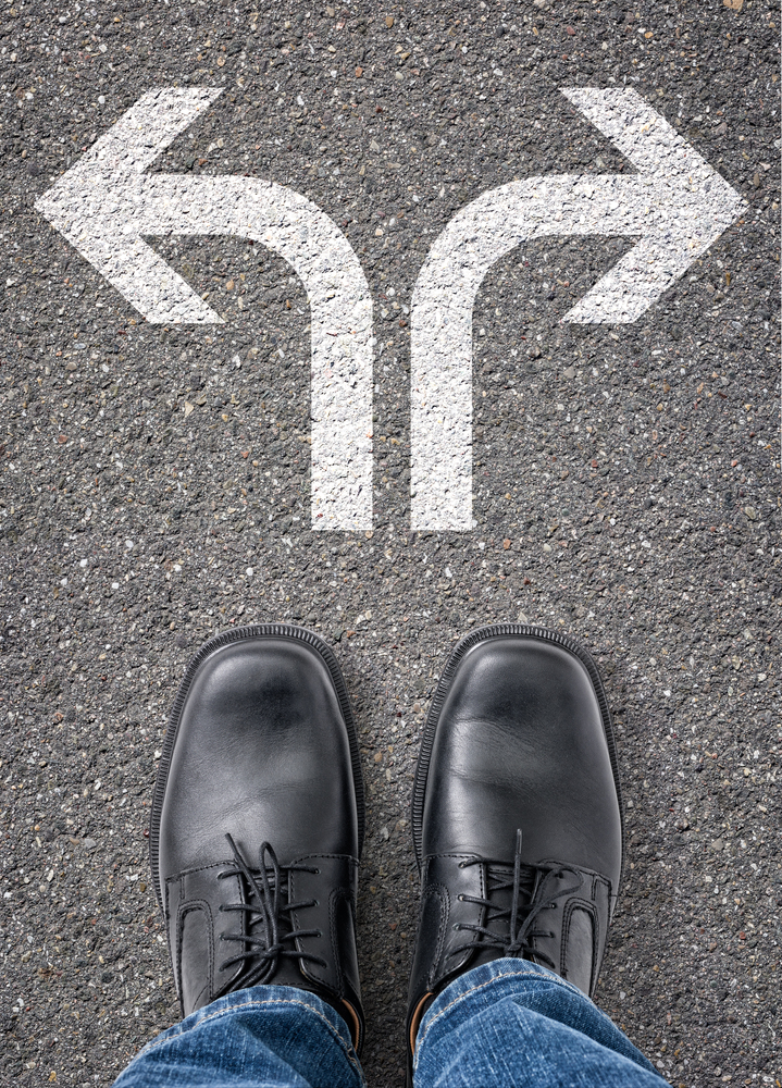 A person wearing black shoes and jeans stands at a fork in the road, with white arrows painted on the pavement pointing left and right, symbolising a choice or decision.