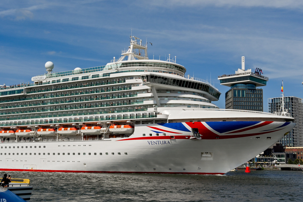 A large white cruise ship named Ventura with a Union Jack design on the bow sails near a city waterfront, with modern buildings and a blue sky in the background.