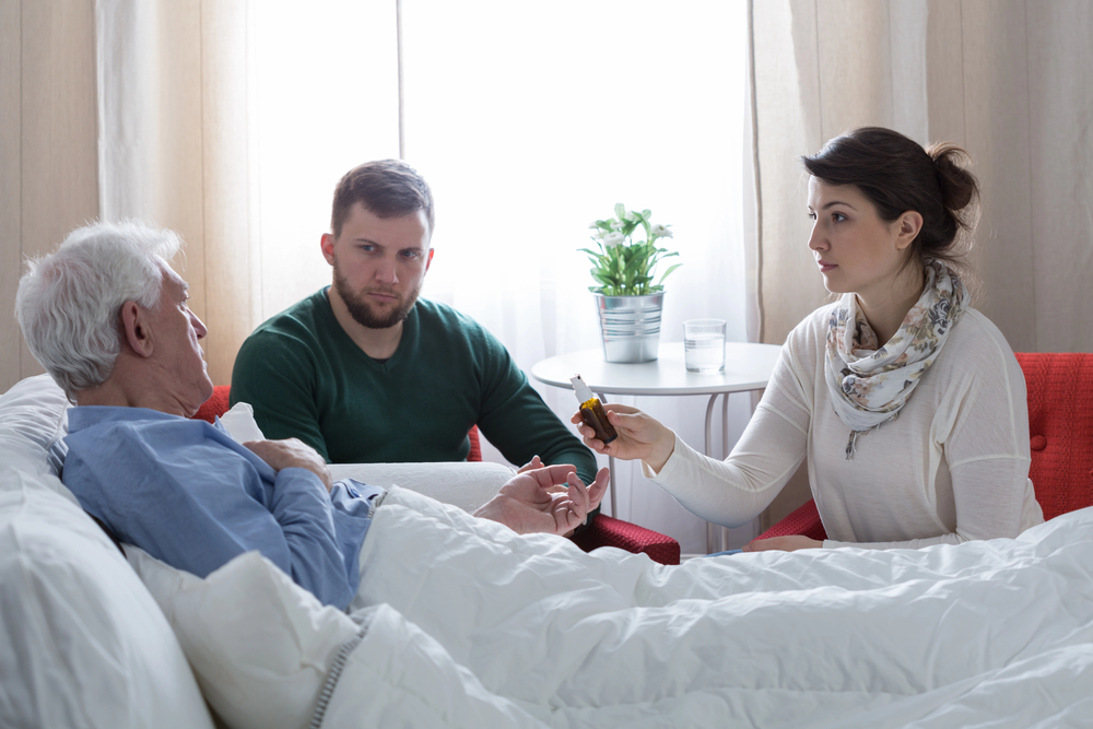 A woman hands medicine to an elderly man lying in bed while a younger man sits beside them, all appearing concerned in a bright, homely room with a plant on a round table in the background.