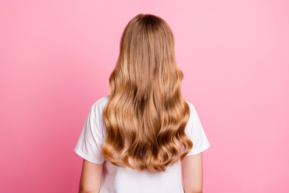 Person with long, wavy, light brown hair wearing a white T-shirt, shown from behind, standing against a solid pink background.