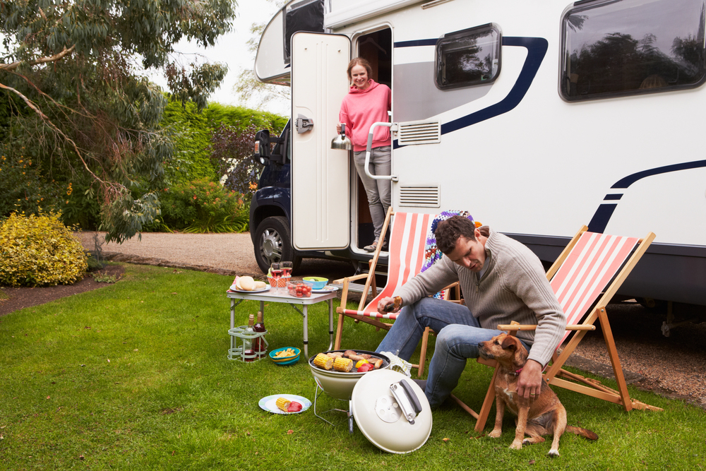 A man sits in a deckchair stroking a small dog while barbecuing food beside a campervan. A woman stands in the camper doorway, and food and drinks are arranged on a table on the grass.