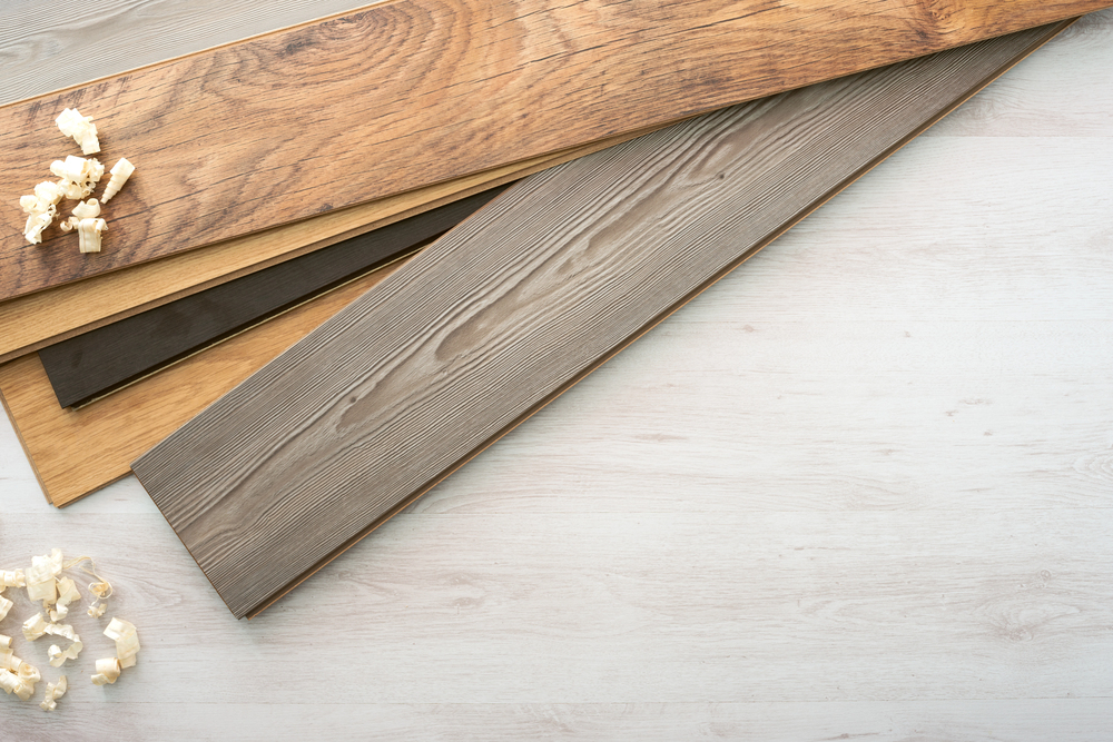 Three wooden laminate floor planks in different shades—brown, dark brown, and grey—are laid out on a light wood surface with wood shavings in the corners of the image.
