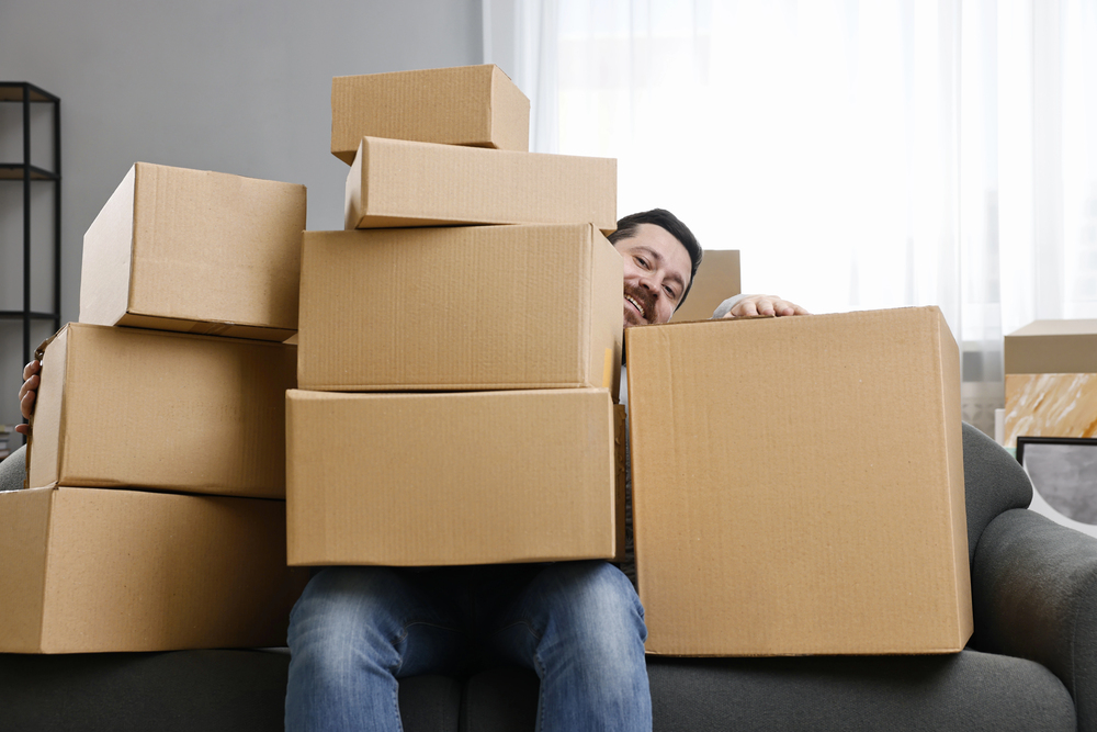 A person sitting on a sofa is mostly hidden behind a large stack of cardboard boxes, with only part of their smiling face visible among the boxes.