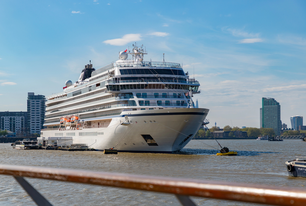 A large white cruise ship is moored on a river near a city, with modern buildings in the background under a clear blue sky. A small tugboat is positioned at the front of the cruise ship.