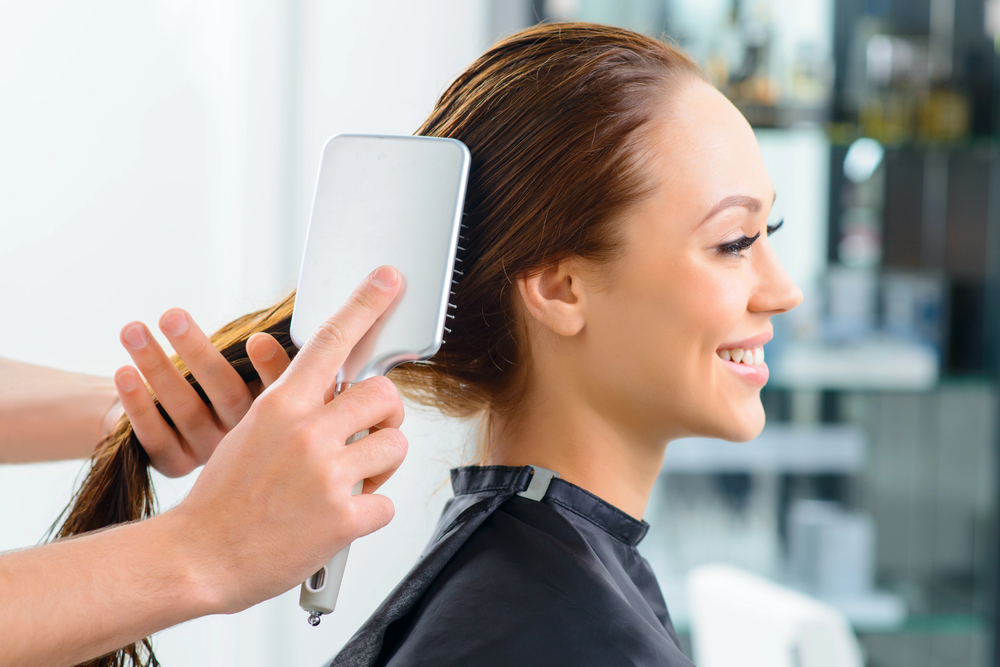 A hairdresser brushes a woman’s long, wet hair in a salon. The woman is smiling and wearing a black salon gown, while the hairdresser uses a white paddle brush. The background shows a blurred salon interior.